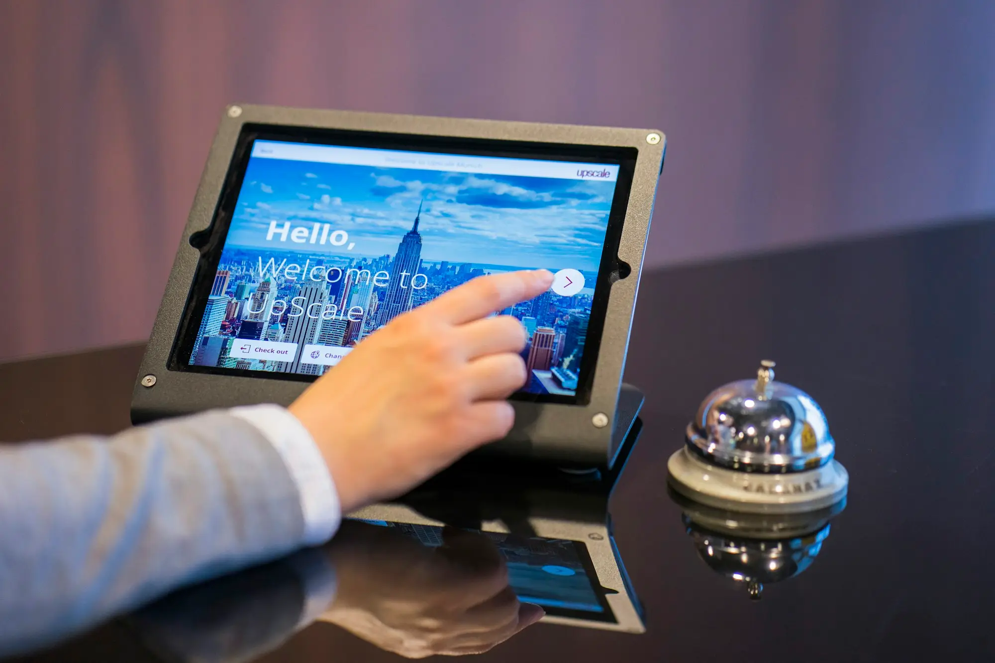 A hand tapping a tablet on a counter beside a silver service bell, waiting for a response